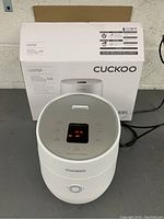 Front view of Cuckoo electric rice cooker powered on with box behind it.