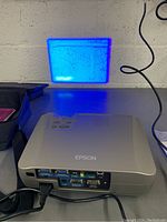 Photo showing the Epson PowerLite projector from the rear with various input ports and projection of a blue image on the wall.