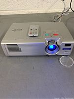 Front view of Epson PowerLite projector with remote on top, powered on showing blue lens light.