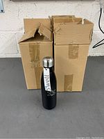 Two images showing the black aluminum S'well water bottles with stainless steel lids in front of two cardboard boxes.