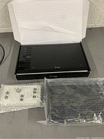 Black box electronic device in open box, white remote control and Netgear router wrapped in plastic and bubble wrap on gray surface.