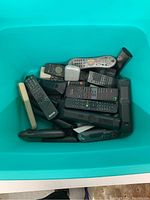 Overview of a teal bin filled with approximately 15 assorted remote controllers of various shapes, sizes, and conditions, including black, gray, and silver colors.