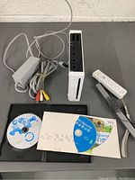 Photo showing Nintendo Wii console standing upright with power indicator on, power adapter, AV cables, Wii Remote with wrist strap, sensor bar, and two game discs including Wii Sports and Mario Kart Wii.