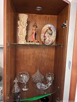 Two shelves showing various decorative figurines, crystal glassware, plate, and vase within a wooden cabinet.