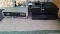 Three devices side by side on tile floor: silver Realistic cassette deck on left, black Pioneer DVD player on top right, and black Panasonic VHS player below the DVD player