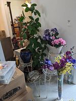 Wide photo showing tall faux tree in woven basket, dark blue ceramic planter, white planter with faux flowers, glass vase with floral stems, and multiple plant stands.