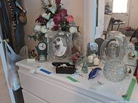 View of assembled lot contents placed on white dresser including glass frames, vases, clock, candle holders, and shoe clips.