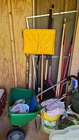 Photo showing various garden tools including yellow metal scoop, rakes, pitchfork, and a green wheelbarrow filled with plastic pipe and bags, inside a wooden shed.