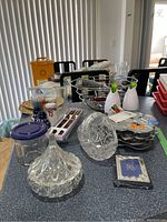Photo showing a variety of kitchenware items such as cut glass salt and pepper shakers, candy dishes, trivets, chopsticks in a box, lighters, and photo albums on a table.