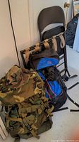 Front and side view showing all four items: US military camo backpack, large travel backpack in black and blue, black padded metal folding chair, and luggage rack with beige fabric straps.