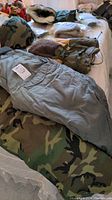 View showing camouflage pants and light olive padded pants with attached belt on bed with other items.