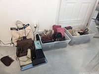 Photo of multiple handbags and purses in two clear plastic bins and laid out on the floor, showing variety of colors and styles.