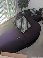 Serpentine style sectional sofa in a living room setting near vertical blinds. Shown is the full length of the curved sofa with several cushions.
