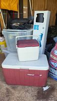 Photo shows a large red and white Coleman cooler (48 qt), smaller cream and red Coleman cooler, two clear plastic tubs stacked, one with a cracked lid, a boxed exercise mat, and the air mattress in black carry bag behind.