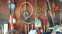 Photo showing a variety of rakes hung on a garage wall, orange Grasshog trimmer, coils of wire and extension cords, some wooden boards and metal rods