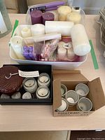 Overview of assorted candles in plastic bins and boxes including pillar, tapered, votive, and tealight candles, with several small glass votive holders.