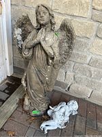 Photo showing full view of the cement angel sculpture standing upright and the smaller ceramic angel figurine on the ground in front of it against a brick wall.