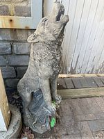 Front view of the cement wolf sculpture showing the full figure sitting and howling, placed on ground near a stone wall and wooden post.