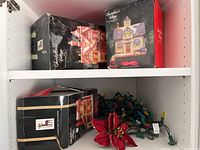 Shelf with boxed Santakins Village porcelain houses, string of colored Christmas lights, and red poinsettia flower decoration.