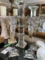 Photo of four Mikasa crystal candle holders on a glass surface with mirror background; shows two distinct styles: three tall with leaf-shaped flared tops and one shorter block-style holder.