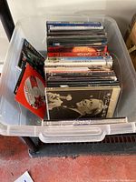 Top-down view of a clear plastic storage bin filled with assorted DVDs, some in jewel cases and some in standard DVD cases. Visible red DVD separate from the jewel cases, bin appears translucent with some label marks.