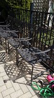 Four black metal outdoor chairs arranged in a row outside near a black metal fence, showing lattice seats, Greek key design backrests, and ornate curved armrests.