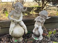 Pair of plastic garden cherubs sitting outdoors on soil with background of garden bed and foliage, showing weathered surface and outdoor use