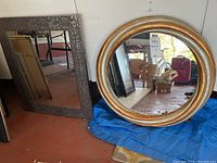 Photo showing both mirrors side by side on the floor. Rectangular mirror has a textured metallic frame; round mirror has a thick gold-tone frame.
