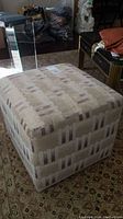 Overall view of a square fabric ottoman showing the geometric upholstery and wear condition.