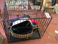 Petmate black wire dog cage containing a woven basket dog bed, dog clothes, and accessories. Cage size approximately 12x19x21 inches.