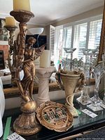 Full view showing the angel candle holder with candle, white pedestal vase, beige two-handled vase, and decorative plate in front.