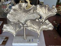 Front view of metal ginkgo leaf sculpture on white stone base showing the detailed veining and silver finish.