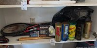 Full view of tennis rackets, pool cue pieces, multiple cans and sleeves of tennis balls on shelf.