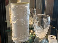 Photo showing both vases on a glass shelf, white ceramic Shafford vase with fish relief on left, clear etched glass vase on right.