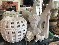 White ceramic decor items including cat sculpture, two elephants, bust, giraffes, and white lattice basket on black table with reflective surface