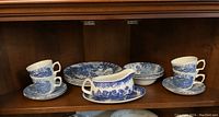 Full view of blue and white Ridgway ironstone cups and saucers, dinner plates, cereal bowls, side plates, and Woodsware Willow pattern sauce boat with saucer on a wooden shelf.