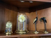 Shelf displaying two anniversary clocks, one with glass dome and one with plastic dome, alongside two metal crane bird figurines in black and brass colors.