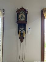 Full view of large oak wall clock with brass pendulums and hanging chains mounted on wall.