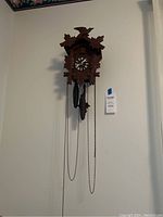 Front view of wooden cuckoo clock with carved leaf and bird motif, showing dial and pinecone weights