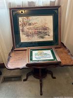 Side table with scalloped edge holding large and small framed landscape artworks with dark green matting