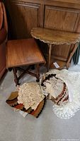 Photo of two wooden tables; demi lune table and rectangular drop-leaf table. Assorted crocheted tableware displayed on floor.