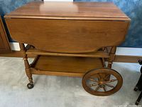 Full view of the tea cart showing the drop leaf sides folded down, large rear wooden wheel, and front caster wheels.