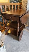 Front view of wooden tea wagon showing one drop leaf raised, drawer closed, turned legs and wooden wheels.