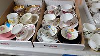 Two cardboard trays each containing six cups and saucers showing assorted floral designs with gold trim