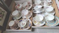Two cardboard boxes each holding six teacup and saucer sets, assorted patterns
