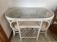 White oval wicker bistro table with glass top and 2 matching wicker chairs tucked underneath. Beige seat cushions visible on the chairs. Table shows wear including paint chipping.