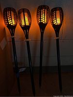 Four black solar-powered outdoor flame effect lights 30 inches tall placed side by side, lit and emitting warm amber glow