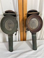 Pair of antique carriage buggy oil lamps from the side showing body, glass, and base with aged metal surfaces.