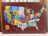 Full frontal view of the metal wall art showing the USA map made from colorful license plate segments on a dark red background.