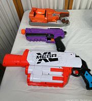 Three Nerf toy blasters arranged on table; white and orange Nerf Mega, purple and black blaster, orange Fortnite-themed blaster